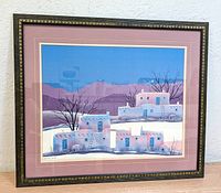 Framed Southwest lithograph print showing traditional adobe buildings in winter with a pink and cream double mat and dark decorative wooden frame.