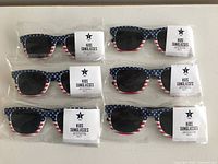 Photo of 8 pairs of kids sunglasses in original packaging, all featuring American flag design on frames.