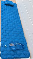 Blue self inflating sleeping pad fully inflated with matching pillow and carry bag on floor beside a white wall.