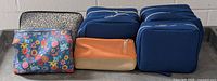 Lot of 8 toiletry bags including 5 blue Nivea bags, 1 floral patterned makeup bag, 1 orange travel bag, and 1 black and white Holt Renew bag arranged on table.