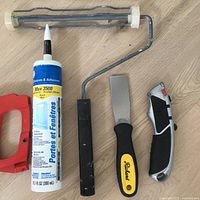 Close-up photo of tools laid out showing window sealant tube, paint roller, putty knife, utility knife, and part of hacksaw handle.