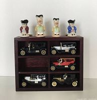 Front view of the wooden display box with five vintage diecast model cars and four miniature ceramic Toby jugs on top, showing missing car compartment and details of jugs and cars.