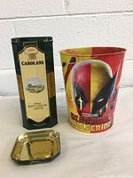 Front view of the Carolans Irish Cream tin can next to the Marvel Deadpool Wolverine popcorn bucket with the tin lid removed and placed in front.
