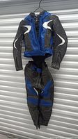 Photo of black, blue, and white Frank Thomas leather motorcycle jacket and matching pants hanging on a hanger, front view.