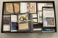Box containing assorted Magic The Gathering cards in various conditions, some bundled with rubber bands, showing multiple cards face up including creature and enchantment types.