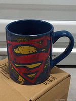 Side view of blue ceramic Superman mug showing distressed logo on cardboard box.