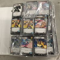 Photo showing a collection of Transformers Trading Card Game cards in plastic sleeves including Armored Plating, Backup Beam, Attack Drone, Bolt of Lightning, and others.