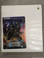 White binder with a clear plastic sleeve on the front containing a Transformers card labeled 'Trypticon Assault Base'.