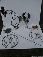 Photo showing all five metal ornaments arranged on a white background and grass: butterfly, frog, rooster with colorful painted sections, cat, and second rooster.
