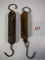 Pair of antique mechanical fish scales shown side by side, one dark heavily worn and one brass-colored with clear measurement markings and hooks.