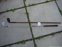 Both golf clubs laid side by side on concrete tiles showing contrast between vintage wooden club and modern metal club.