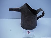 Front left angled view of antique metal oil can showing handle, spout and cylindrical body with patina and rust.
