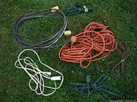 Top-down view of assorted coiled extension cords laid on grass showing multiple colors including black, orange, white, green, and brown cords with varying plug types.