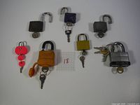 Top-down photo showing seven assorted padlocks with keys arranged on white surface with a grid paper marked '18'.