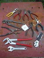Full view of the assorted pliers and wrenches laid out on a rusted metal surface. Various sizes and handle colors visible.