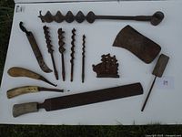 Photo shows entire collection of vintage hand tools including auger bits, axe head, hand plane, rasp, sickle, and cast iron piece arranged on white background.