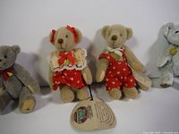 Close-up photo of three miniature teddy bears, one grey with bowtie, two beige wearing red outfit with heart patterns, with original tags visible