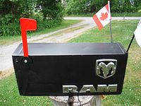 Side view of a rectangular black handmade mailbox with chrome Dodge Ram emblem and RAM letters, red mailbox flag raised and Canadian flag attached at the back.