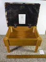 Photo of foot stool lid open showing wooden interior storage compartment, underside of lid with black fabric and label.