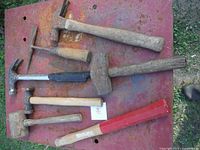 Six hammers and mallets laid out on a rusty red metal surface, showing various wooden and metal handles and signs of wear.