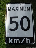 Photo of front of large rectangular metal traffic sign, displaying 'MAXIMUM 50 km/h' in black letters on white reflective background with black border.