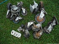Image showing a scattered lot of 26 casters including metal casters with some rust and a set of white plastic rollers on grass.
