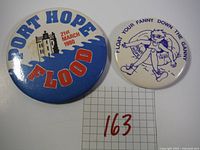 Top view of two pin-back buttons with front graphics, one large blue/red Port Hope Flood 1980 button and one smaller white FYFDTG 1982 button
