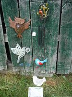 Four garden decor bird items displayed against green wooden fence: metal owl wall hanging, white cutout bird on spike, blue and red bird decoration, and small white ceramic bird figurine on ground.