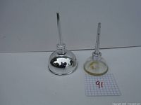 Two vintage oil cans displayed side by side on a white surface. One is chrome finish and the other has a transparent amber base.