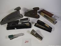Seven vintage staplers arranged on a white surface with a label '203' grid paper for lot identification. Includes various sizes and styles, some metal and some with plastic parts. One has a wooden top.
