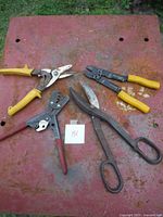 Four hand tools laid on a rusty red surface: yellow-handled wire crimper, red-handled cutters, large heavy-duty metal scissors, yellow-handled wire snips.