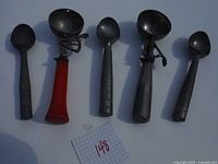 Five vintage metal ice cream scoops, two with red handles and three solid metal with ribbed handles, arranged on a neutral background.