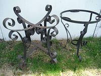 Wide angle view of two ornate metal table bases/plant stands outside on grass and near a white wall.