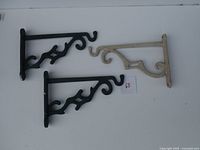 Three aluminum plant hangers (two black, one light grey) laid flat on white surface with a small numbered paper