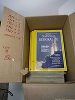Box containing stacks of National Geographic magazines with visible yellow borders; one cover shows 'Columbia' space shuttle launch.