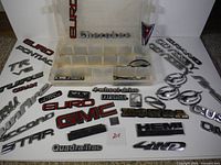 Top down view of assorted vintage vehicle badges and emblems laid out, including Pontiac, Cherokee, Euro, GMC logos, 4 wheel drive, Quadra-Trac script, and more.