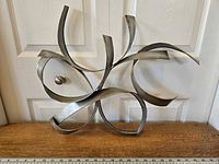 Front view of abstract metal wall art placed on a wooden surface in front of a white door, showing flowing curved metal strips and round accent piece.