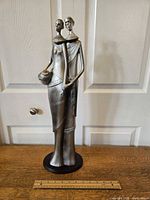 Front angled view of tall metallic sculpture depicting an African tribal couple with a ruler for scale showing 7 inches width and 22.25 inches height.