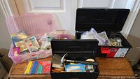 Photo showing two black plastic tool/storage boxes and one purple plastic storage box on wooden table, filled with assorted tools and hardware
