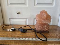 Side view of lamp showing Buddha carving, electrical cord with switch, and wooden base with ruler for scale.
