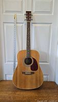 Full front view of the guitar showing dreadnought body shape, natural wood finish, and white trim; standing next to a tape measure for scale.