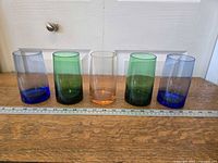 Five drinking glasses in blue, green, and orange colors on a wooden surface next to a ruler showing approximately 5.5 inches height each.