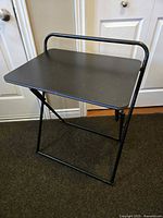 Full view of black folding table standing open, showing tabletop and tubular metal frame with handle.
