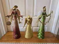 Photo showing three hollow metal angels with glittery surfaces in front of a white door. Left angel is purple and holding ornaments, center angel smaller gold with wings, right angel tall green with star decorations.