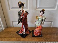 Pair of two geisha girl figurines standing on a wooden surface in front of a white door, shown with a measuring ruler for scale.