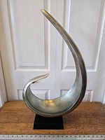 Front view of wave sculpture showing curved shape and silver finish, placed on wooden surface with ruler in front.