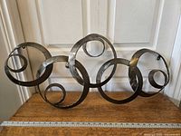 Front view of metal wall art with continuous large and small circle rings, placed on wooden surface with ruler showing size.