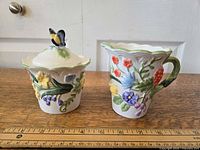 Milk container with floral relief decoration and butterfly lid handle, displayed on wooden surface with ruler for size reference.