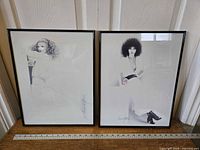Two framed black and white prints featuring female portraits by Sara Moon, titled 'Rebecca' and 'Catherine II', positioned side by side on the floor against a wall.
