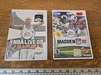 Front view of both Nintendo Wii game cases: NBA Live 09 All-Play and Madden NFL 10 displayed side by side on wooden surface with measuring tape below.